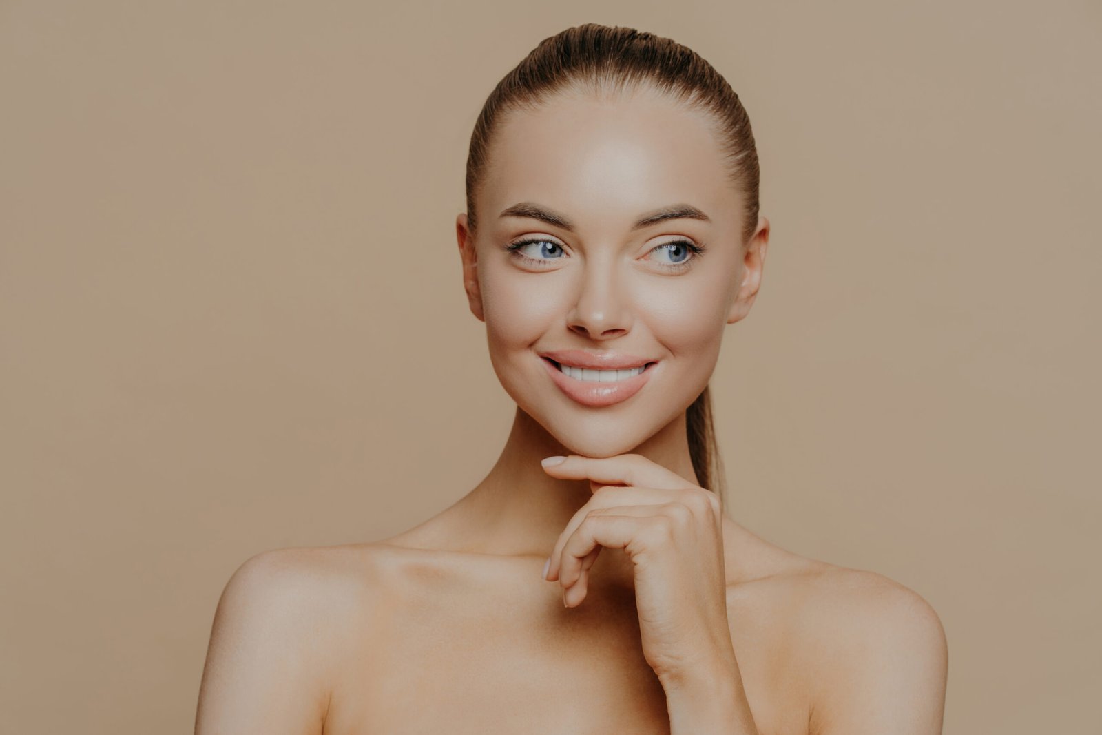 smiling well groomed young woman with beauty face care, healthy glowing skin, touches chin gently and looks aside, poses shirtless against brown studio background, loves her skin, feels refreshed